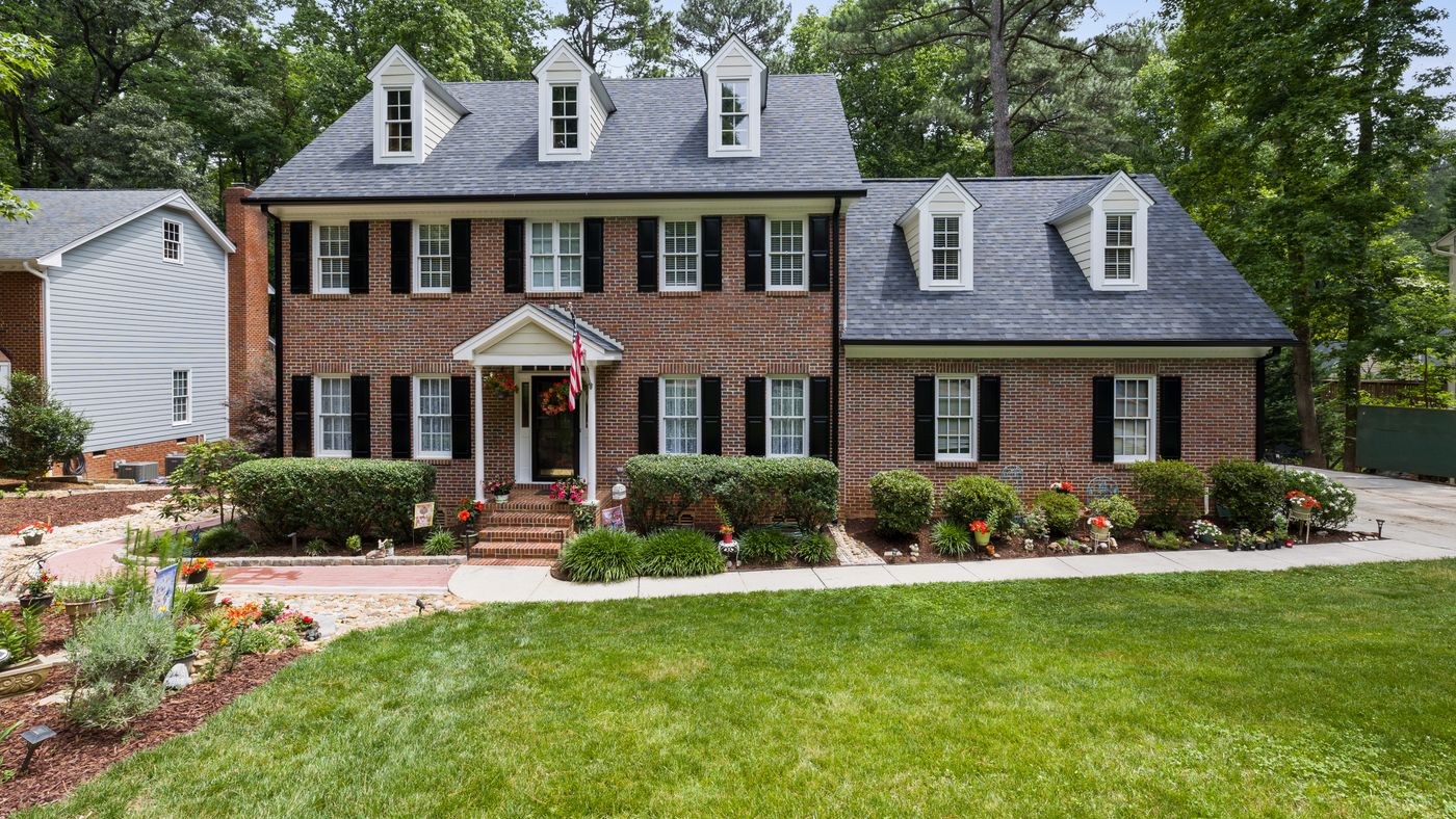 Brick colonial house with dormer windows and a mown front lawn on a wooded street