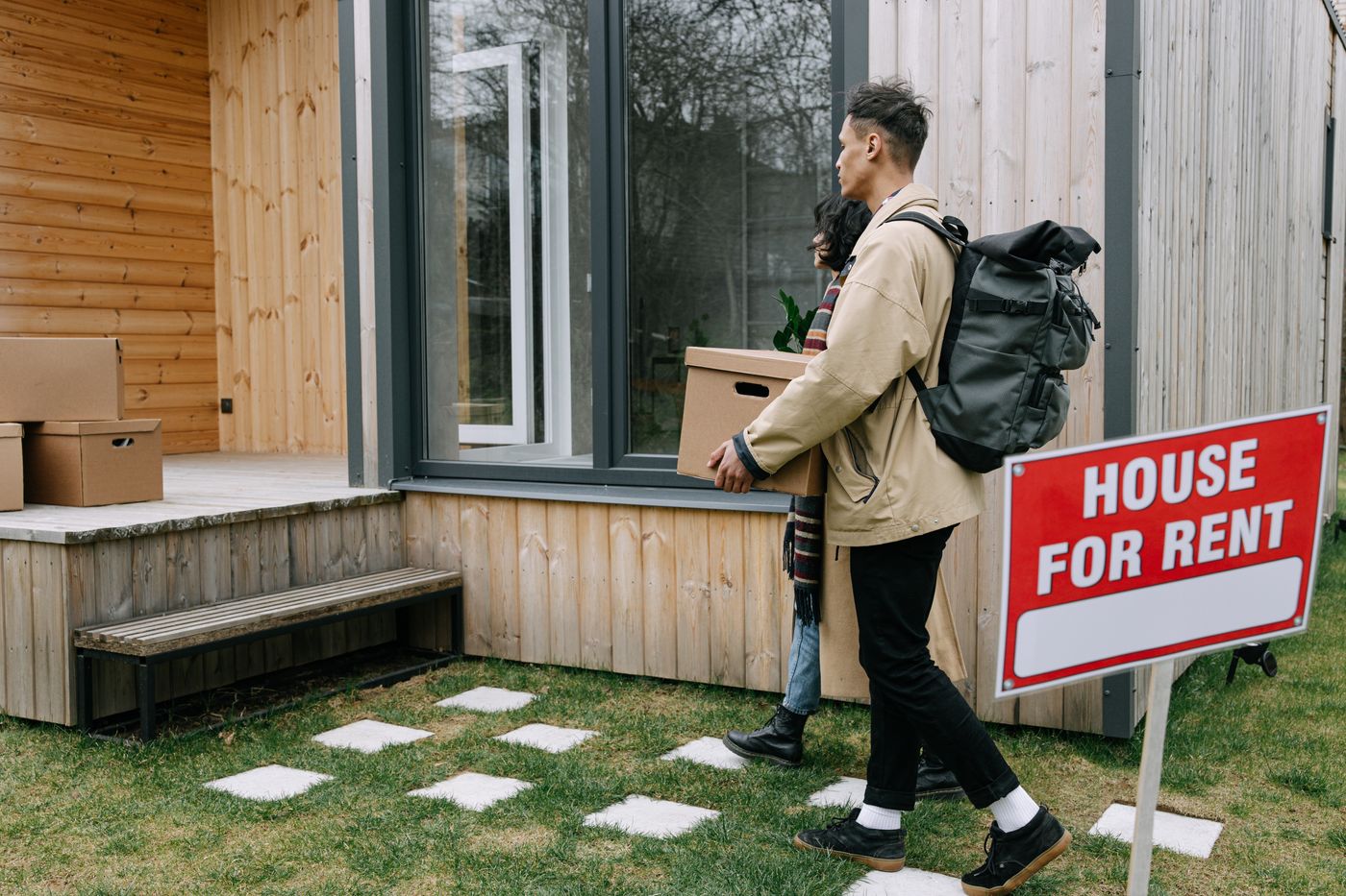 A tenant carrying moving boxes into a rental home beside a house-for-rent sign